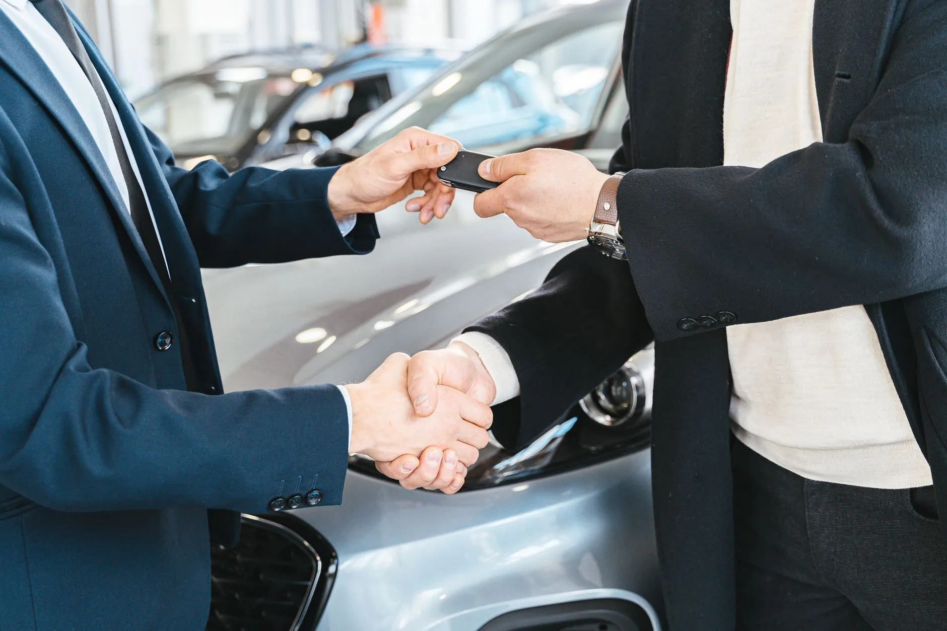 Vendedor entrega a chave do carro ao comprador com um aperto de mão, ao lado de um carro prata